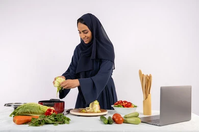 تعلم مهارة الطهي وإعداد الطعام عبر الحاسب المحمول، ربة منزل سعودية تقوم بتحضير وصفة بالخضراوات الطازجة، احترافية اختيار الخضار، بورتريه لسيدة عربية خليجية اماراتية محجبة ترتدي عباءة تحضر وجبة صحية مفيدة، مشاركة الوصفات على مواقع الانترنت، خلفية بيضاء
