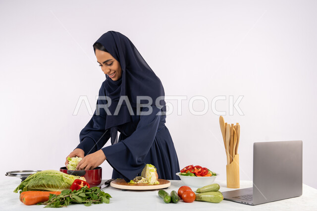 مشاركة الوصفات على مواقع الانترنت، تعلم مهارة الطهي وإعداد الطعام عبر الحاسب المحمول، ربة منزل سعودية تقوم بتحضير وصفة بالخضراوات الطازجة، احترافية اختيار الخضار، بورتريه لسيدة عربية خليجية اماراتية محجبة ترتدي عباءة تحضر وجبة صحية مفيدة، خلفية بيضاء