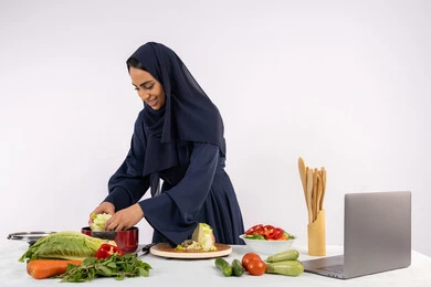 Sharing recipes on websites, learning the skill of cooking and preparing food via laptop, a Saudi housewife preparing a recipe with fresh vegetables, professionalism in choosing vegetables, a portrait of a veiled Emirati Gulf Arab woman wearing an abaya preparing a healthy and useful meal, white background