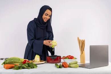 تعلم مهارة الطهي وإعداد الطعام عبر الحاسب المحمول، ربة منزل سعودية تقوم بتحضير وصفة بالخضراوات الطازجة، احترافية اختيار الخضار، بورتريه لسيدة عربية خليجية اماراتية محجبة ترتدي عباءة تحضر وجبة صحية مفيدة، مشاركة الوصفات على مواقع الانترنت، خلفية بيضاء