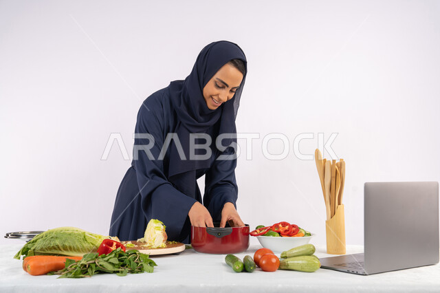 مشاركة الوصفات على مواقع الانترنت، تعلم مهارة الطهي وإعداد الطعام عبر الحاسب المحمول، ربة منزل سعودية تقوم بتحضير وصفة بالخضراوات الطازجة، احترافية اختيار الخضار، بورتريه لسيدة عربية خليجية اماراتية محجبة ترتدي عباءة تحضر وجبة صحية مفيدة، خلفية بيضاء