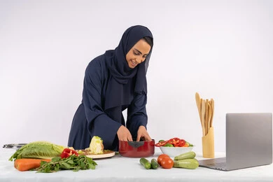 مشاركة الوصفات على مواقع الانترنت، تعلم مهارة الطهي وإعداد الطعام عبر الحاسب المحمول، ربة منزل سعودية تقوم بتحضير وصفة بالخضراوات الطازجة، احترافية اختيار الخضار، بورتريه لسيدة عربية خليجية اماراتية محجبة ترتدي عباءة تحضر وجبة صحية مفيدة، خلفية بيضاء