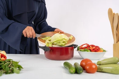 إعداد الطعام الصحي للأسرة، حب تعلم مهارة الطهي، ربة منزل سعودية تقوم بتحضير وصفة بالخضراوات الطازجة، احترافية اختيار الخضار، بورتريه مقرب لسيدة عربية خليجية اماراتية ترتدي عباءة تحضر وجبة صحية مفيدة، خلفية بيضاء