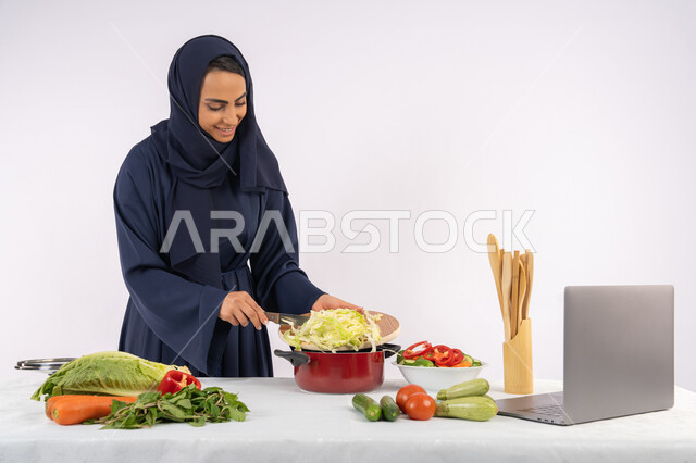 الاستمتاع بإعداد الطعام الصحي، تعلم مهارة الطهي عبر الحاسب المحمول، ربة منزل سعودية تقوم بتحضير وصفة بالخضراوات الطازجة، احترافية اختيار الخضار، بورتريه لسيدة عربية خليجية اماراتية محجبة ترتدي عباءة تحضر وجبة صحية مفيدة، مشاركة الوصفات على مواقع الانترنت، خلفية بيضاء