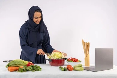 الاستمتاع بإعداد الطعام الصحي، تعلم مهارة الطهي عبر الحاسب المحمول، ربة منزل سعودية تقوم بتحضير وصفة بالخضراوات الطازجة، احترافية اختيار الخضار، بورتريه لسيدة عربية خليجية اماراتية محجبة ترتدي عباءة تحضر وجبة صحية مفيدة، مشاركة الوصفات على مواقع الانترنت، خلفية بيضاء