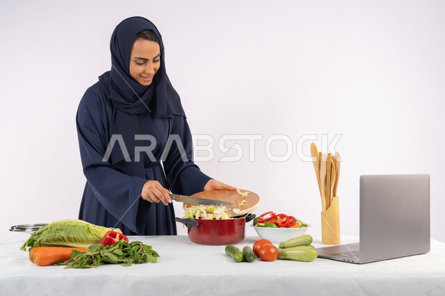 نشر ومشاركة الوصفات على مواقع الانترنت، تعلم مهارة الطهي وإعداد الطعام عبر الحاسب المحمول، ربة منزل سعودية تقوم بتحضير وصفة بالخضراوات الطازجة، احترافية اختيار الخضار، بورتريه لسيدة عربية خليجية اماراتية محجبة ترتدي عباءة تحضر وجبة صحية مفيدة، خلفية بيضاء