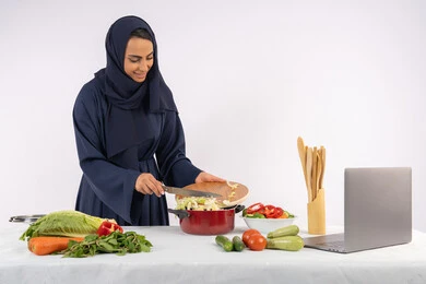 نشر ومشاركة الوصفات على مواقع الانترنت، تعلم مهارة الطهي وإعداد الطعام عبر الحاسب المحمول، ربة منزل سعودية تقوم بتحضير وصفة بالخضراوات الطازجة، احترافية اختيار الخضار، بورتريه لسيدة عربية خليجية اماراتية محجبة ترتدي عباءة تحضر وجبة صحية مفيدة، خلفية بيضاء