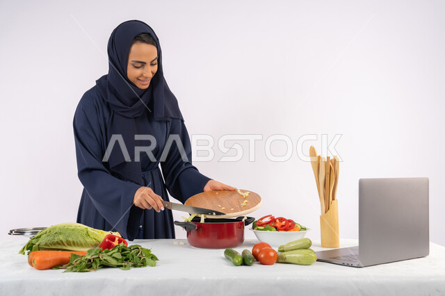 تعلم مهارة الطهي وإعداد الطعام عبر الحاسب المحمول، ربة منزل سعودية تقوم بتحضير وصفة بالخضراوات الطازجة، احترافية اختيار الخضار، بورتريه لسيدة عربية خليجية اماراتية محجبة ترتدي عباءة تحضر وجبة صحية مفيدة، مشاركة الوصفات على مواقع الانترنت، خلفية بيضاء