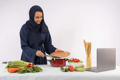 تعلم مهارة الطهي وإعداد الطعام عبر الحاسب المحمول، ربة منزل سعودية تقوم بتحضير وصفة بالخضراوات الطازجة، احترافية اختيار الخضار، بورتريه لسيدة عربية خليجية اماراتية محجبة ترتدي عباءة تحضر وجبة صحية مفيدة، مشاركة الوصفات على مواقع الانترنت، خلفية بيضاء