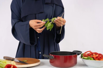 شغف التدرب على مهارة الطهي وإعداد الطعام، احترافية اختيار الخضار، ربة منزل سعودية تقوم بتحضير سلطة الخضراوات الطازجة، بورتريه مقرب لسيدة عربية خليجية اماراتية ترتدي عباءة تحضر طبق صحي مفيد، خلفية بيضاء