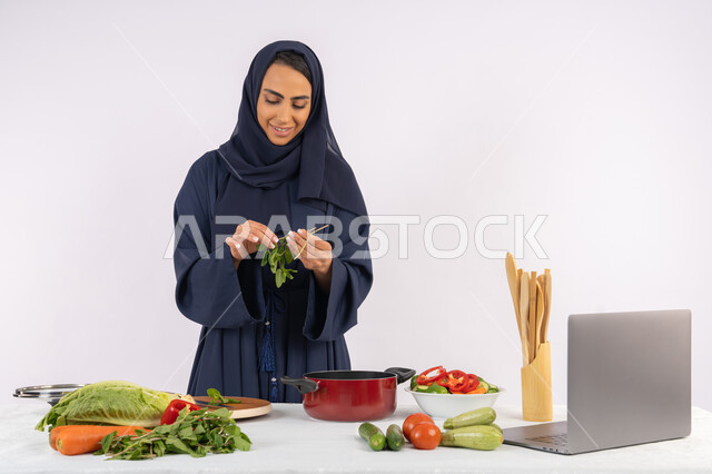 شغف التدرب على مهارة الطهي وإعداد الطعام، احترافية اختيار الخضار، مشاركة الوصفات على مواقع الانترنت عبر الحاسوب المحمول، ربة منزل سعودية تقوم بتحضير سلطة الخضراوات الطازجة، بورتريه لسيدة عربية خليجية اماراتية محجبة ترتدي عباءة تحضر طبق صحي مفيد، خلفية بيضاء