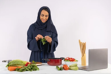 شغف التدرب على مهارة الطهي وإعداد الطعام، احترافية اختيار الخضار، مشاركة الوصفات على مواقع الانترنت عبر الحاسوب المحمول، ربة منزل سعودية تقوم بتحضير سلطة الخضراوات الطازجة، بورتريه لسيدة عربية خليجية اماراتية محجبة ترتدي عباءة تحضر طبق صحي مفيد، خلفية بيضاء