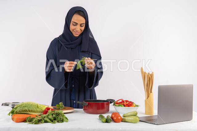احترافية اختيار الخضار، مشاركة الوصفات على مواقع الانترنت عبر الحاسوب المحمول، ربة منزل سعودية تقوم بتحضير سلطة الخضراوات الطازجة، شغف التدرب على مهارة الطهي وإعداد الطعام، بورتريه لسيدة عربية خليجية اماراتية محجبة ترتدي عباءة تحضر طبق صحي مفيد، خلفية بيضاء