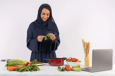 احترافية اختيار الخضار، مشاركة الوصفات على مواقع الانترنت عبر الحاسوب المحمول، ربة منزل سعودية تقوم بتحضير سلطة الخضراوات الطازجة، شغف التدرب على مهارة الطهي وإعداد الطعام، بورتريه لسيدة عربية خليجية اماراتية محجبة ترتدي عباءة تحضر طبق صحي مفيد، خلفية بيضاء