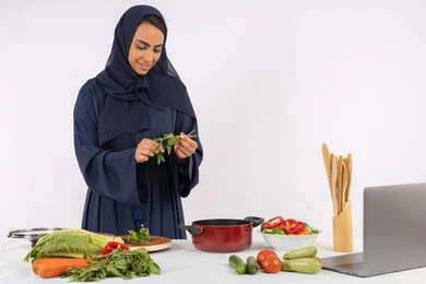 شغف التدرب على مهارة الطهي وإعداد الطعام، احترافية اختيار الخضار، مشاركة الوصفات على مواقع الانترنت عبر الحاسوب المحمول، ربة منزل سعودية تقوم بتحضير سلطة الخضراوات الطازجة، بورتريه لسيدة عربية خليجية اماراتية محجبة ترتدي عباءة تحضر طبق صحي مفيد، خلفية بيضاء