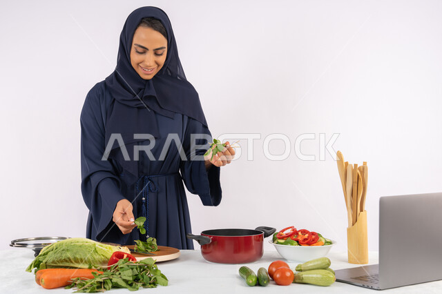 المتعة أثناء تحضير الطعام الصحي، احترافية اختيار الخضار، مشاركة الوصفات على الانترنت عبر الحاسوب ، ربة منزل سعودية تقوم بتحضير سلطة الخضراوات الطازجة، شغف التدرب على مهارة الطهي، بورتريه لسيدة عربية خليجية اماراتية محجبة ترتدي عباءة تحضر طبق صحي مفيد، خلفية بيضاء