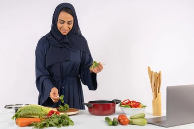 المتعة أثناء تحضير الطعام الصحي، احترافية اختيار الخضار، مشاركة الوصفات على الانترنت عبر الحاسوب ، ربة منزل سعودية تقوم بتحضير سلطة الخضراوات الطازجة، شغف التدرب على مهارة الطهي، بورتريه لسيدة عربية خليجية اماراتية محجبة ترتدي عباءة تحضر طبق صحي مفيد، خلفية بيضاء