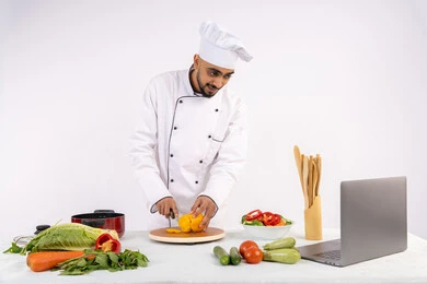 تعلم مهارة الطهي وأعداد الطعام عبر الحاسب المحمول، بورتريه لرجل عربي خليجي اماراتي يرتدي زي العمل يقوم بتحضير وصفة طبق صحي مفيد من الانترنت، شيف سعودي يقوم بتقطيع الخضراوات الطازجة، استخدام المكونات والمقادير المناسبة للوجبة، خلفية بيضاء