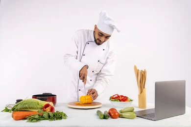 المتعة بتعلم مهارة الطهي وأعداد الطعام عبر الحاسب المحمول، بورتريه لرجل عربي خليجي اماراتي يرتدي زي العمل يقوم بتحضير وصفة طبق صحي مفيد من الانترنت، شيف سعودي يقوم بتقطيع الخضراوات الطازجة، استخدام المكونات والمقادير المناسبة للوجبة، خلفية بيضاء