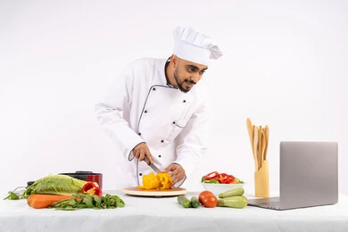 تعلم مهارة الطهي وأعداد الطعام عبر الحاسب المحمول، بورتريه لرجل عربي خليجي اماراتي يرتدي زي العمل يقوم بتحضير وصفة طبق صحي مفيد من الانترنت، شيف سعودي يقوم بتقطيع الخضراوات الطازجة، استخدام المكونات والمقادير المناسبة للوجبة، خلفية بيضاء