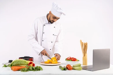 Learning cooking and food preparation skills via laptop, portrait of an Emirati Gulf Arab man wearing work clothes preparing a healthy and useful dish recipe from the internet, a Saudi chef chopping fresh vegetables, using the appropriate ingredients and quantities for the meal, white background