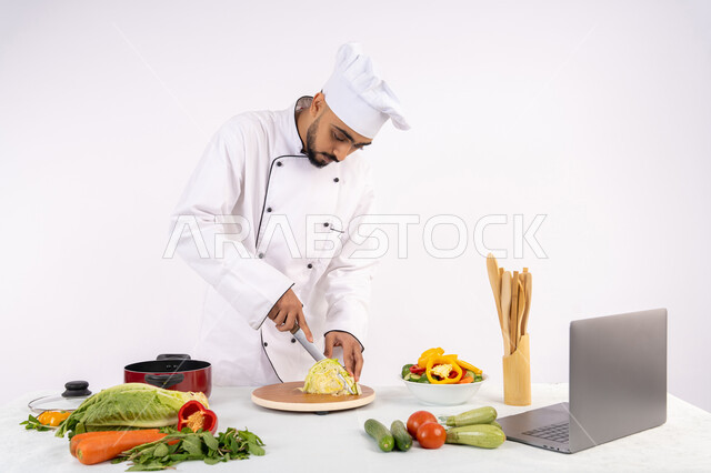 التدرب على مهارة الطهي وأعداد الطعام عبر الحاسب المحمول، بورتريه لرجل عربي خليجي اماراتي يرتدي زي العمل يقوم بتحضير وصفة طبق صحي مفيد من الانترنت، شيف سعودي يقوم بتقطيع الخضراوات الطازجة، استخدام المكونات والمقادير المناسبة للوجبة، خلفية بيضاء