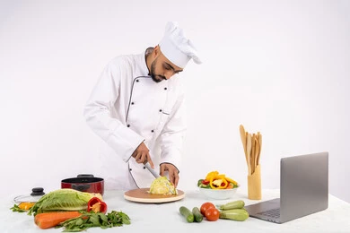 Practicing cooking and food preparation skills via laptop, portrait of an Emirati Gulf Arab man wearing work clothes preparing a healthy dish recipe from the internet, a Saudi chef chopping fresh vegetables, using appropriate ingredients and quantities for the meal, white background