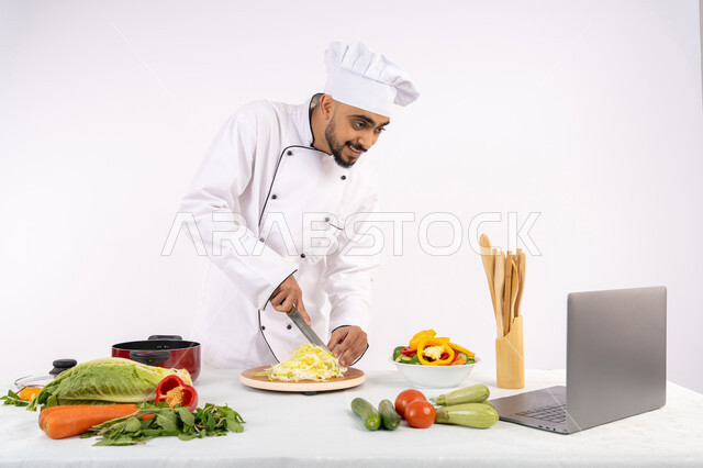 التدرب على مهارة الطهي وأعداد الطعام عبر الحاسب المحمول، بورتريه لرجل عربي خليجي اماراتي يرتدي زي العمل يقوم بتحضير وصفة طبق صحي مفيد من الانترنت، شيف سعودي يقوم بتقطيع الخضراوات الطازجة، استخدام المكونات والمقادير المناسبة للوجبة، خلفية بيضاء