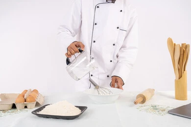 طاهي سعودي يقوم بتجهيز المكونات لصناعة الخبز والفطور، إعداد المخبوزات والمعجنات الشهية، بورتريه مقرب لرجل عربي خليجي اماراتي يرتدي زي العمل يقوم بخفق المكونات مع الدقيق الابيض لتحضير الطعام، خلفية بيضاء