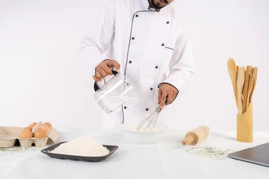 إعداد المخبوزات والمعجنات الشهية، بورتريه مقرب لرجل عربي خليجي اماراتي يرتدي زي العمل يقوم بخفق المكونات مع الدقيق الابيض لتحضير الطعام، شيف سعودي يقوم بتجهيز المكونات لصناعة الخبز والفطور، خلفية بيضاء