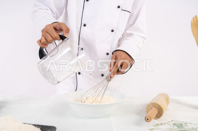 طاهي سعودي يقوم بتجهيز المكونات لصناعة الخبز والفطور، إعداد المخبوزات والمعجنات الشهية، بورتريه مقرب لرجل عربي خليجي اماراتي يرتدي زي العمل يقوم بخفق المكونات مع الدقيق الابيض لتحضير الطعام، خلفية بيضاء