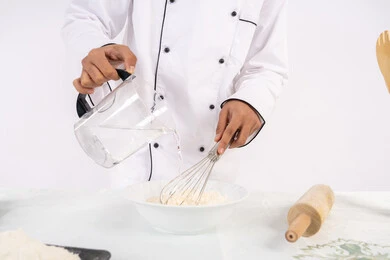 طاهي سعودي يقوم بتجهيز المكونات لصناعة الخبز والفطور، إعداد المخبوزات والمعجنات الشهية، بورتريه مقرب لرجل عربي خليجي اماراتي يرتدي زي العمل يقوم بخفق المكونات مع الدقيق الابيض لتحضير الطعام، خلفية بيضاء