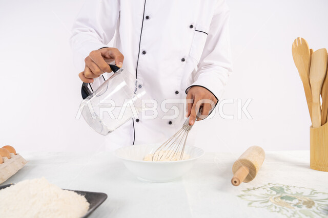 Preparing delicious baked goods and pastries. Close-up portrait of an Emirati Gulf Arab man in work clothes mixing ingredients with white flour to prepare food. A Saudi chef prepares ingredients for making bread and breakfast. White background.