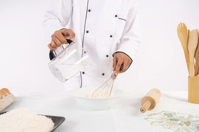Preparing delicious baked goods and pastries. Close-up portrait of an Emirati Gulf Arab man in work clothes mixing ingredients with white flour to prepare food. A Saudi chef prepares ingredients for making bread and breakfast. White background.