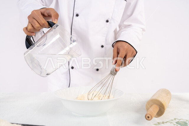 Preparing delicious baked goods and pastries. Close-up portrait of an Emirati Gulf Arab man in work clothes mixing ingredients with white flour to prepare food. A Saudi chef prepares ingredients for making bread and breakfast. Whiteط background.