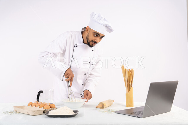 تصوير ومشاركة الوصفة عبر المواقع باستخدام الحاسوب المحمول، طاهي سعودي يقوم بتجهيز المكونات لصناعة الخبز والفطور، إعداد المخبوزات والمعجنات الشهية، بورتريه لرجل عربي خليجي اماراتي يرتدي زي العمل يقوم بخفق المكونات مع الدقيق الابيض لتحضير الطعام، خلفية بيضاء