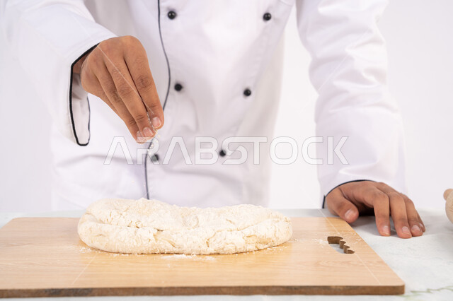 A Saudi chef prepares ingredients for making bread and breakfast, preparing delicious baked goods and pastries, a portrait of an Arab Gulf Emirati man wearing work clothes sprinkling white flour on the dough to prepare food, white background