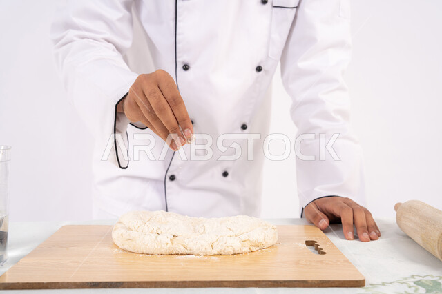 A Saudi chef prepares ingredients for making bread and breakfast, preparing delicious baked goods and pastries, a portrait of an Arab Gulf Emirati man wearing work clothes sprinkling white flour on the dough to prepare food, white background