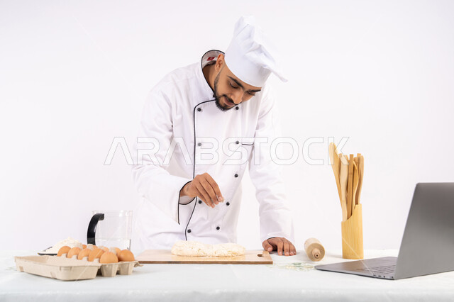 مشاركة الوصفة عبر المواقع باستخدام الحاسوب المحمول، تحضير وإعداد المخبوزات والمعجنات الشهية، بورتريه لرجل عربي خليجي اماراتي يرتدي زي العمل يقوم بفرد العجين باستخدام الشوبك (النشابة)، شيف سعودي يقوم بتجهيز المكونات لصناعة الخبز والفطور، خلفية بيضاء