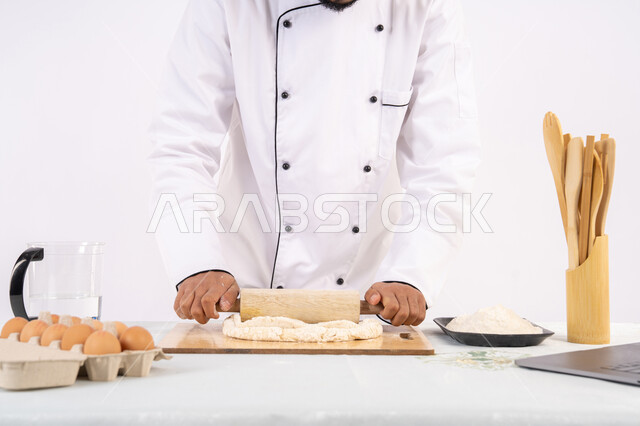 Sharing the recipe across websites using a laptop, preparing and baking delicious baked goods and pastries, portrait of an Emirati Gulf Arab man wearing work clothes rolling out dough using a rolling pin, a Saudi chef preparing ingredients for making bread and breakfast, white background
