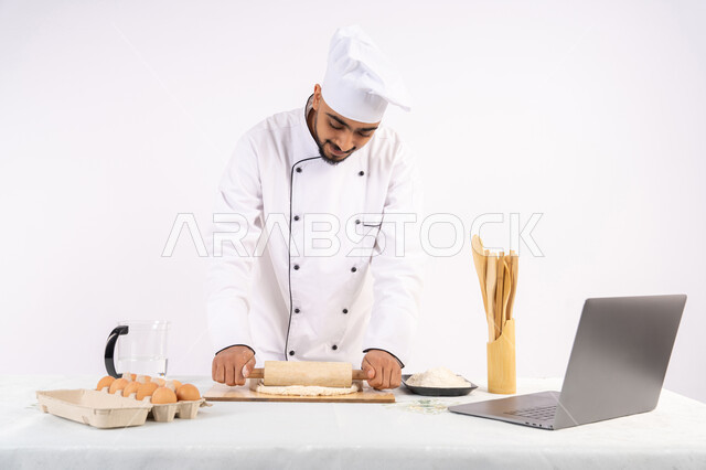 مشاركة الوصفة عبر المواقع باستخدام الحاسوب المحمول، تحضير وإعداد المخبوزات والمعجنات الشهية، بورتريه لرجل عربي خليجي اماراتي يرتدي زي العمل يقوم بفرد العجين باستخدام الشوبك (النشابة)، شيف سعودي يقوم بتجهيز المكونات لصناعة الخبز والفطور، خلفية بيضاء
