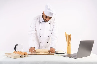 Sharing the recipe across websites using a laptop, preparing and baking delicious baked goods and pastries, portrait of an Emirati Gulf Arab man wearing work clothes rolling out dough using a rolling pin, a Saudi chef preparing ingredients for making bread and breakfast, white background