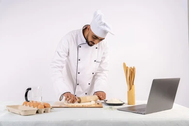 Sharing the recipe across websites using a laptop, preparing and baking delicious baked goods and pastries, portrait of an Emirati Gulf Arab man wearing work clothes rolling out dough using a rolling pin, a Saudi chef preparing ingredients for making bread and breakfast, white background