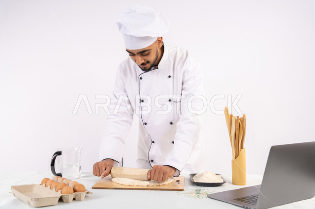 مشاركة الوصفة عبر المواقع باستخدام الحاسوب المحمول، تحضير وإعداد المخبوزات والمعجنات الشهية، بورتريه لرجل عربي خليجي اماراتي يرتدي زي العمل يقوم بفرد العجين باستخدام الشوبك (النشابة)، شيف سعودي يقوم بتجهيز المكونات لصناعة الخبز والفطور، خلفية بيضاء