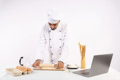 Photographing and sharing the recipe on social media using a laptop, preparing and baking delicious baked goods and pastries, portrait of an Emirati Gulf Arab man wearing work clothes rolling out dough using a rolling pin, Saudi chef preparing ingredients for making bread and breakfast, white background