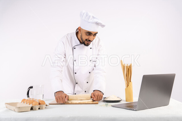 تصوير ومشاركة الوصفة عبر المواقع باستخدام الحاسوب المحمول، تحضير وإعداد المخبوزات والمعجنات الشهية، بورتريه لرجل عربي خليجي اماراتي يرتدي زي العمل يقوم بفرد العجين باستخدام الشوبك (النشابة)، شيف سعودي يقوم بتجهيز المكونات لصناعة الخبز والفطور، خلفية بيضاء
