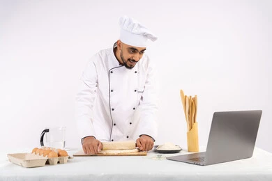 Photographing and sharing the recipe on social media using a laptop, preparing and baking delicious baked goods and pastries, portrait of an Emirati Gulf Arab man wearing work clothes rolling out dough using a rolling pin, Saudi chef preparing ingredients for making bread and breakfast, white background