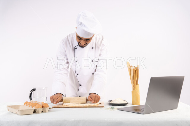 تصوير ومشاركة الوصفة عبر المواقع باستخدام الحاسوب المحمول، تحضير وإعداد المخبوزات والمعجنات الشهية، بورتريه لرجل عربي خليجي اماراتي يرتدي زي العمل يقوم بفرد العجين باستخدام الشوبك (النشابة)، شيف سعودي يقوم بتجهيز المكونات لصناعة الخبز والفطور، خلفية بيضاء