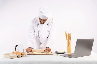 Photographing and sharing the recipe on social media using a laptop, preparing and baking delicious baked goods and pastries, portrait of an Emirati Gulf Arab man wearing work clothes rolling out dough using a rolling pin, Saudi chef preparing ingredients for making bread and breakfast, white background
