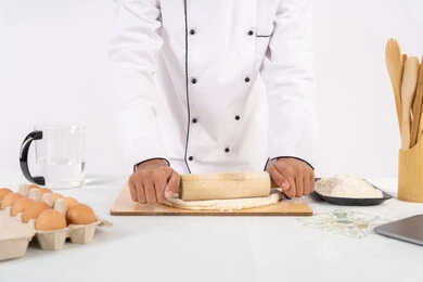 شيف سعودي يقوم بتجهيز المكونات لصناعة الخبز والفطور، تحضير وإعداد المخبوزات والمعجنات الشهية، بورتريه مقرب لرجل عربي خليجي اماراتي يرتدي زي العمل يقوم بفرد العجين باستخدام الشوبك (النشابة)، خلفية بيضاء